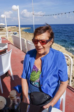 Emekli, güneş gözlüğü takan yaşlı kadın deniz kenarındaki restoranda oturuyor.