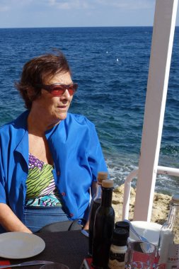 Emekli, güneş gözlüğü takan yaşlı kadın deniz kenarındaki restoranda oturuyor.