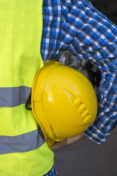 Cropped image of a construction worker wearing a yellow safety vest and keeping a yellow helmet underarm