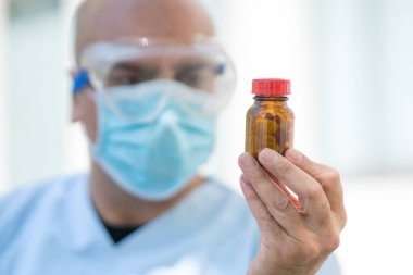 Beyaz önlük, ameliyat maskesi ve tıbbi güvenlik gözlüğü takmış elinde bir kutu hapla odaklanmış bir doktorun portresi. Seçici odak