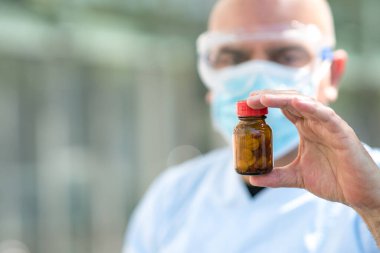 Beyaz önlük, ameliyat maskesi ve tıbbi güvenlik gözlüğü takmış elinde bir kutu hapla odaklanmış bir doktorun portresi. Seçici odak