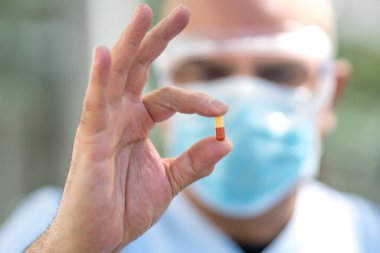 Cerrahi maske takan ve gözlük takan bir doktor parmaklarında ilaç hapı olduğunu gösteriyor.
