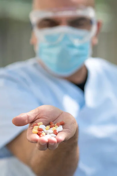 Doctor wearing surgical mask and medical safety goggles holding a handful of medication pills in his hand