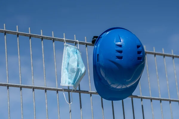 Occupational safety and protection against adverse conditions at work. Blue hard hat and protective surgical mask hanging on scaffolding. Selective focus on foreground