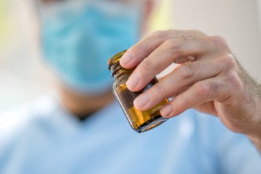 Cerrahi maske takan bir doktor elinde bir şişe tıbbi şurup tutuyor. Önplandaki kavanoza odaklan