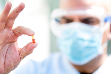 Ameliyat maskesi ve tıbbi güvenlik gözlüğü takan odaklanmış bir doktor parmaklarında ilaç hapları tutuyor.