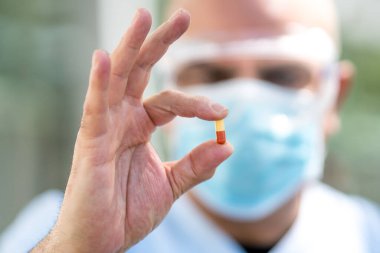 Ameliyat maskesi ve tıbbi güvenlik gözlüğü takan odaklanmış bir doktor parmaklarında ilaç hapları tutuyor.