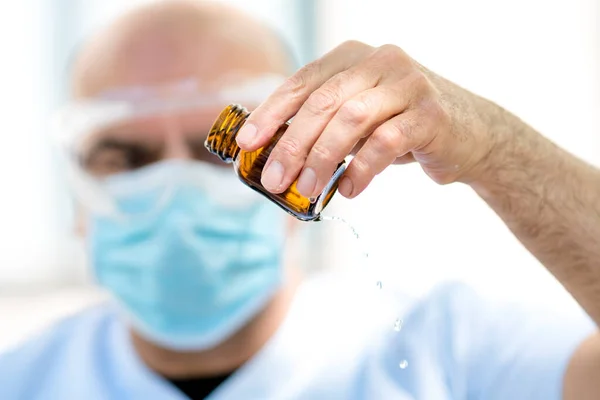 Doctor wearing a surgical mask and medical safety goggles emptying a bottle of expired liquid antibiotics. Selective focus on foreground