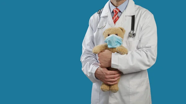 Medical doctor with a stethoscope on shoulders holds a teddy bear in a medical mask.