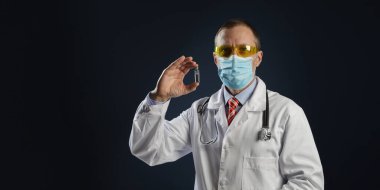 Olgun bir erkek doktor elinde aşı tutuyor. Tıbbi koruyucu maskeli ve gözlüklü doktor..