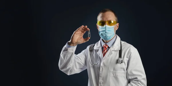 Olgun bir erkek doktor elinde aşı tutuyor. Tıbbi koruyucu maskeli ve gözlüklü doktor..