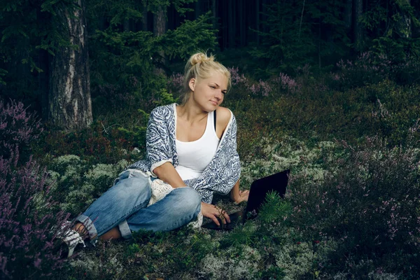 Gyönyörű fiatal nő ül a füvön az erdőben, és keres számítógépet. Nő használ laptopot szabadtéri. — Stock Fotó