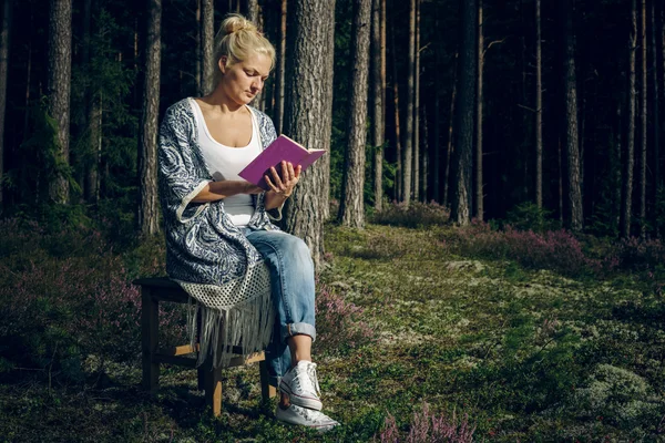 Young beautiful woman sitting on a bench in the woods and reading a book — Stock Fotó