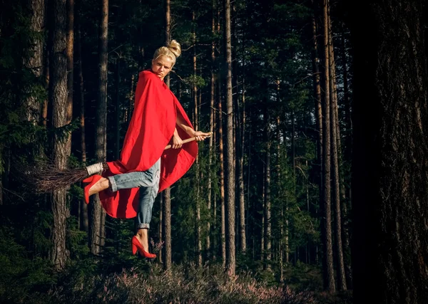 Modern kızgın cadı kot pantolon ve kırmızı pelerin ormanda bir süpürge üzerinde uçan. Levitation. Cadılar Bayramı tatili.