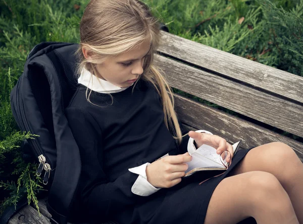 Gadis pirang yang cantik dengan seragam sekolah duduk di bangku taman dan membaca buku . — Stok Foto