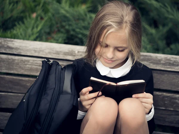 Bella ragazza bionda in uniforme scolastica si siede sulla panchina del parco e libro di lettura . — Foto Stock