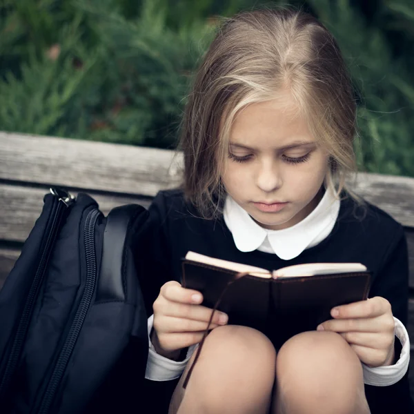 Bella ragazza bionda in uniforme scolastica si siede sulla panchina del parco e libro di lettura . — Foto Stock