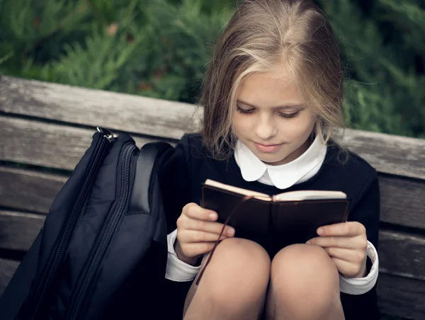 Schönes blondes Mädchen in Schuluniform sitzt auf Parkbank und liest Buch. — Stockfoto