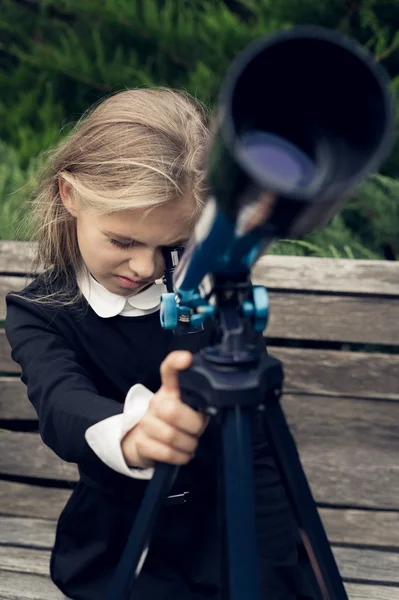 Beautiful blond girl in a school uniform in the park looking through  telescope — Stock Photo, Image