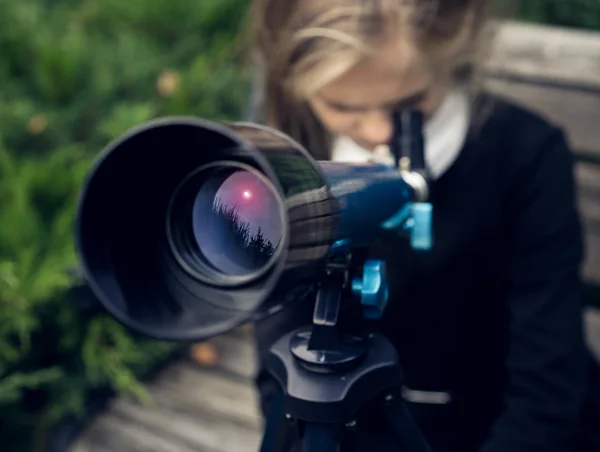 Beautiful blond girl in a school uniform in the park looking through  telescope — Stock Photo, Image