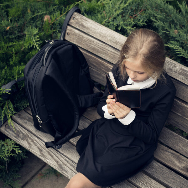 Beautiful blond girl in a school uniform sits on park bench and reading book.
