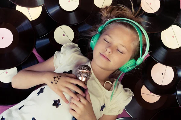 Hermosa chica acostada junto a discos de vinilo sosteniendo un micrófono y escuchando música en los auriculares . — Foto de Stock