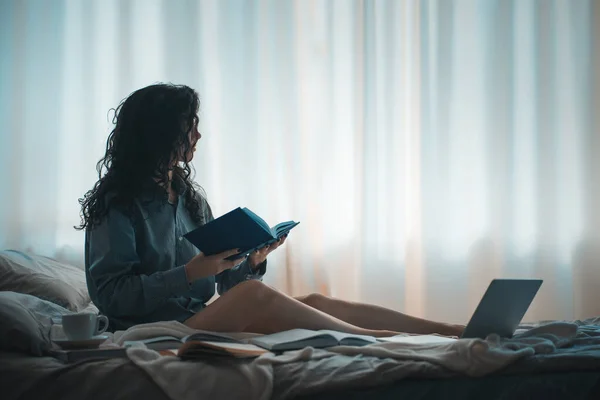 一个漂亮的黑发女人躺在早晨的床上，带着书和咖啡 — 图库照片