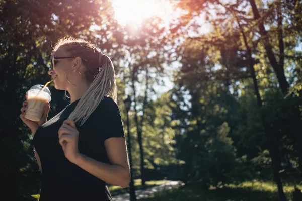 Giovane bella intrecciata femminile bere frullato con caramello nel parco soleggiato — Foto Stock