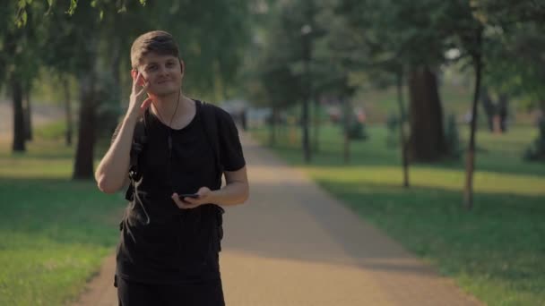 Молодой человек гуляет в парке и слушает музыку — стоковое видео