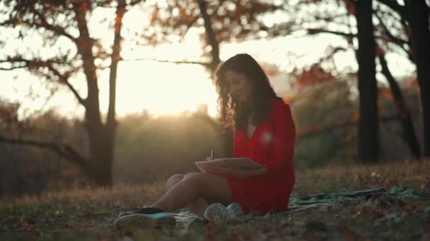 Kaunis nuori nainen kirjoittaa päiväkirjaa syksyllä puistossa — kuvapankkivideo