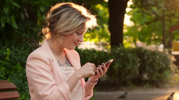 Belle femme blanche blonde utilisant un smartphone dans le parc à la journée ensoleillée — Video