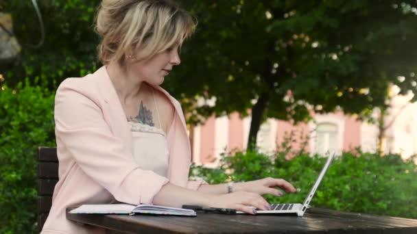 Attractive blonde white woman working freelance with laptop and smartphone in park at sunny day — Stock Video