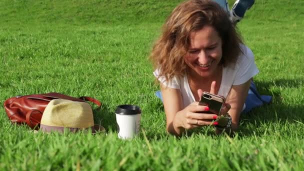 Mujer joven atractiva en el césped utiliza un teléfono inteligente en el día soleado — Vídeos de Stock