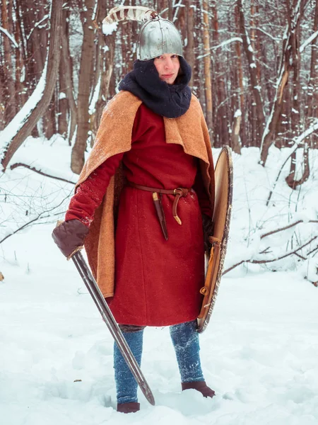 Guerriero franco in costume storico con spada, scudo, casco nella foresta invernale Immagine Stock