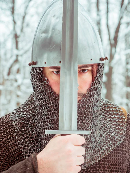 Guerriero vichingo medievale in maglia a catena e casco tiene una spada nella foresta invernale Foto Stock Royalty Free