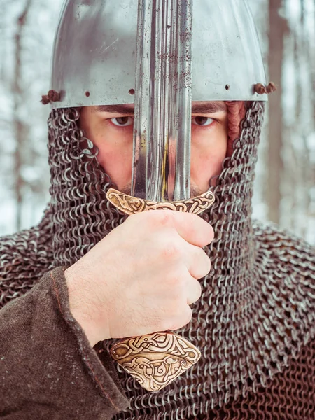 Guerriero vichingo medievale in maglia a catena e casco tiene una spada nella foresta invernale Foto Stock