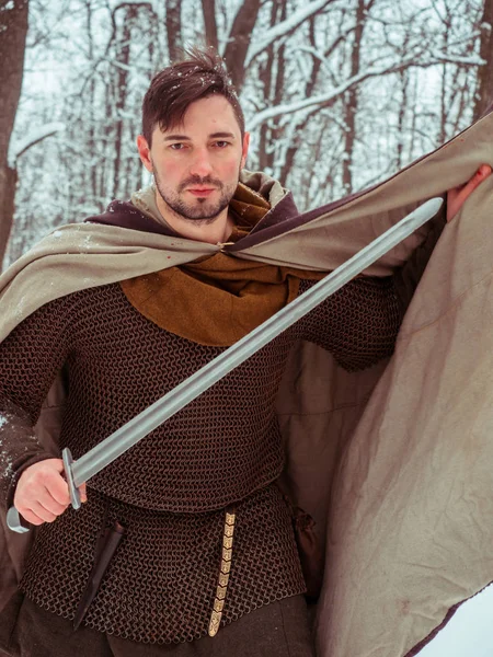 Guerriero vichingo medievale in maglia a catena e casco tiene una spada nella foresta invernale Immagini Stock Royalty Free