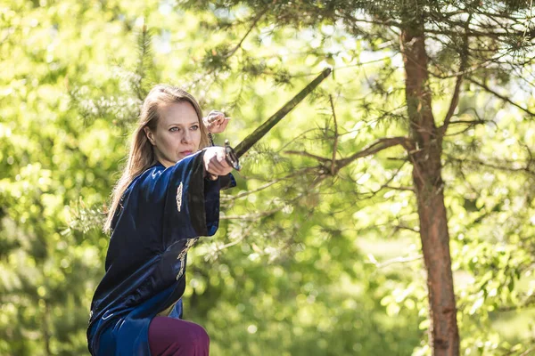 A woman warrior with a sword poses on a stump in the forest. Practice Wushu fencing in nature