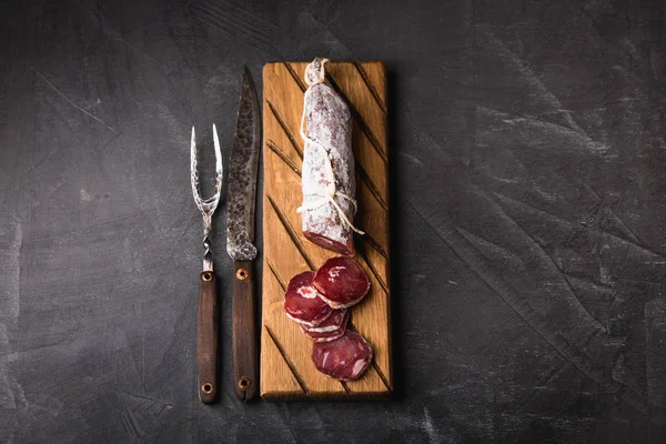 Fresh salami on a vintage Board. Vintage Cutlery fork and knife on a black coated background. Copy space.