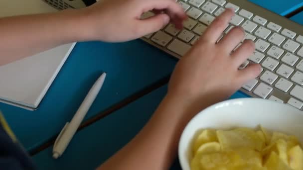 Il ragazzo è seduto al computer a fare i compiti, giocare a un gioco, guardare video e mangiare patatine fritte . — Video Stock