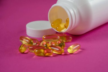 Sarı omega 3 balık yağı kapsülleri mor arka planda beyaz şişeden dökülüyor..