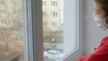 Tıbbi maskeli bir çocuk pencereden dışarı bakıyor. Karantina, koronavirüs, covid 19 'da kendini izole etme..