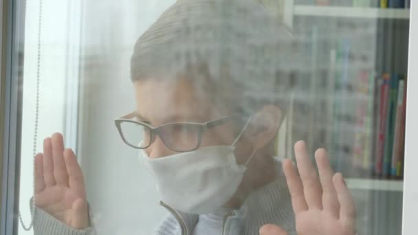 Young boy in a medical mask and glasses looks out the window. Self-isolation in quarantine, coronavirus, covid 19. — Stock Video