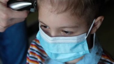 Evdeki berber dükkanında maskeli bir çocuk. Baba, küçük oğlunun saçını kırpıcı ile kesiyor. Coronavirus, karantina, covid19.