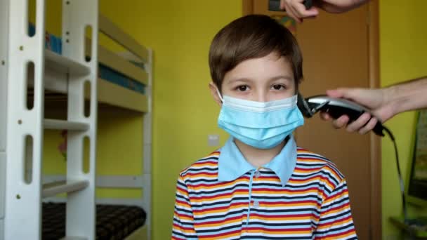 Jovem com máscara médica numa barbearia caseira. Pai cortando cabelo de filho pequeno com clipper. Coronavírus, quarentena, covid19 . — Vídeo de Stock