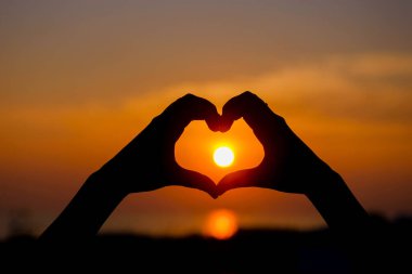 Kadın elleri günbatımı siluetiyle bir kalp şekli oluşturuyor..