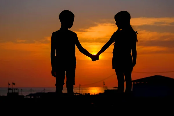 Çocuklar gün batımının arka planında el ele tutuşuyorlar. Genç bir erkek ve kızın siluetleri..