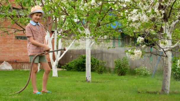 Kleiner Junge mit Hut gießt einen Baum mit einem Schlauch auf der Datscha. — Stockvideo
