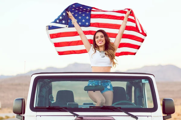 Счастливая молодая девушка, стоящая в кабриолете, держа американский флаг . — стоковое фото