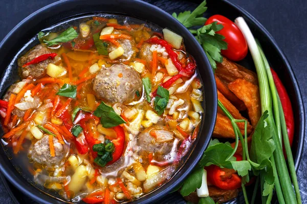 Суп с фрикадельками в черной тарелке, нарезанные овощи - лук, петрушка, перец, помидор на черном фоне — стоковое фото